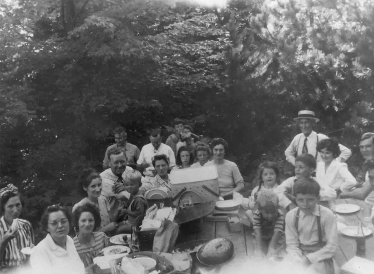 A group of adults and children gathers around a picnic table laden with food in a wooded setting.