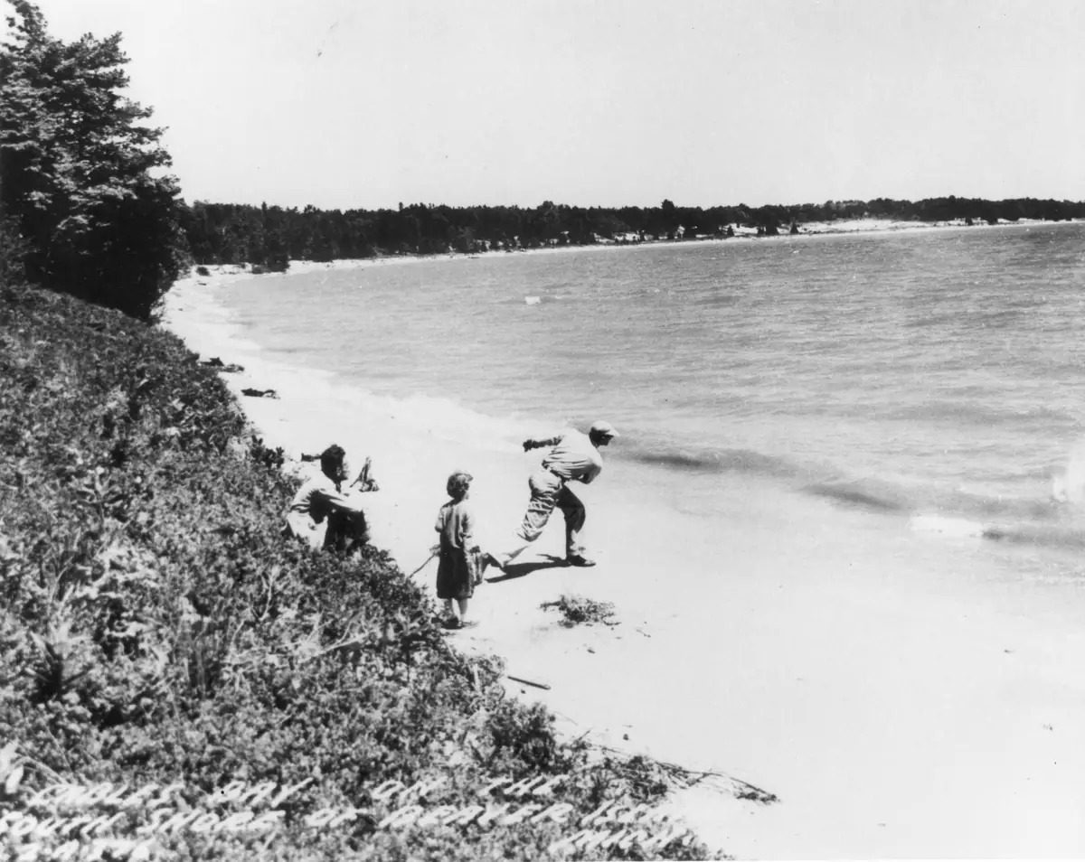 Three people stand and sit on a sandy beach beside calm water and a forested shoreline.