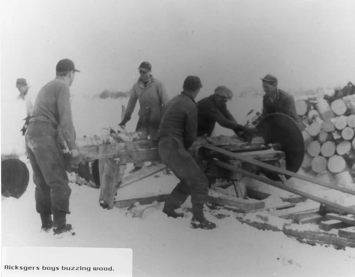 Five people work around a large circular saw in a snowy outdoor lumber yard beside stacked logs.