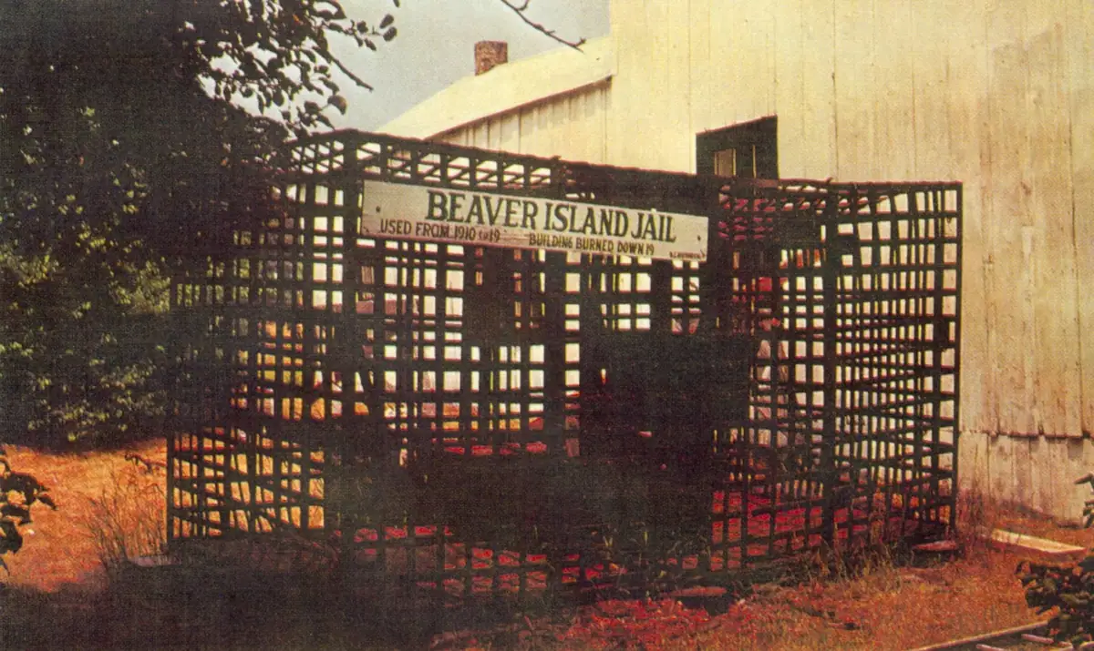 A wooden lattice enclosure stands beside a pale-sided building, with a sign identifying it as a jail.