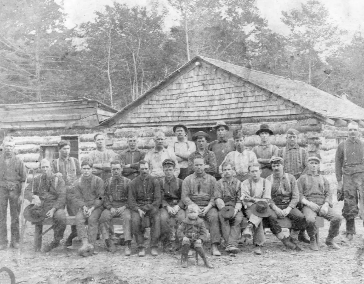 A large group of men and one child poses in front of a log building in a wooded setting.