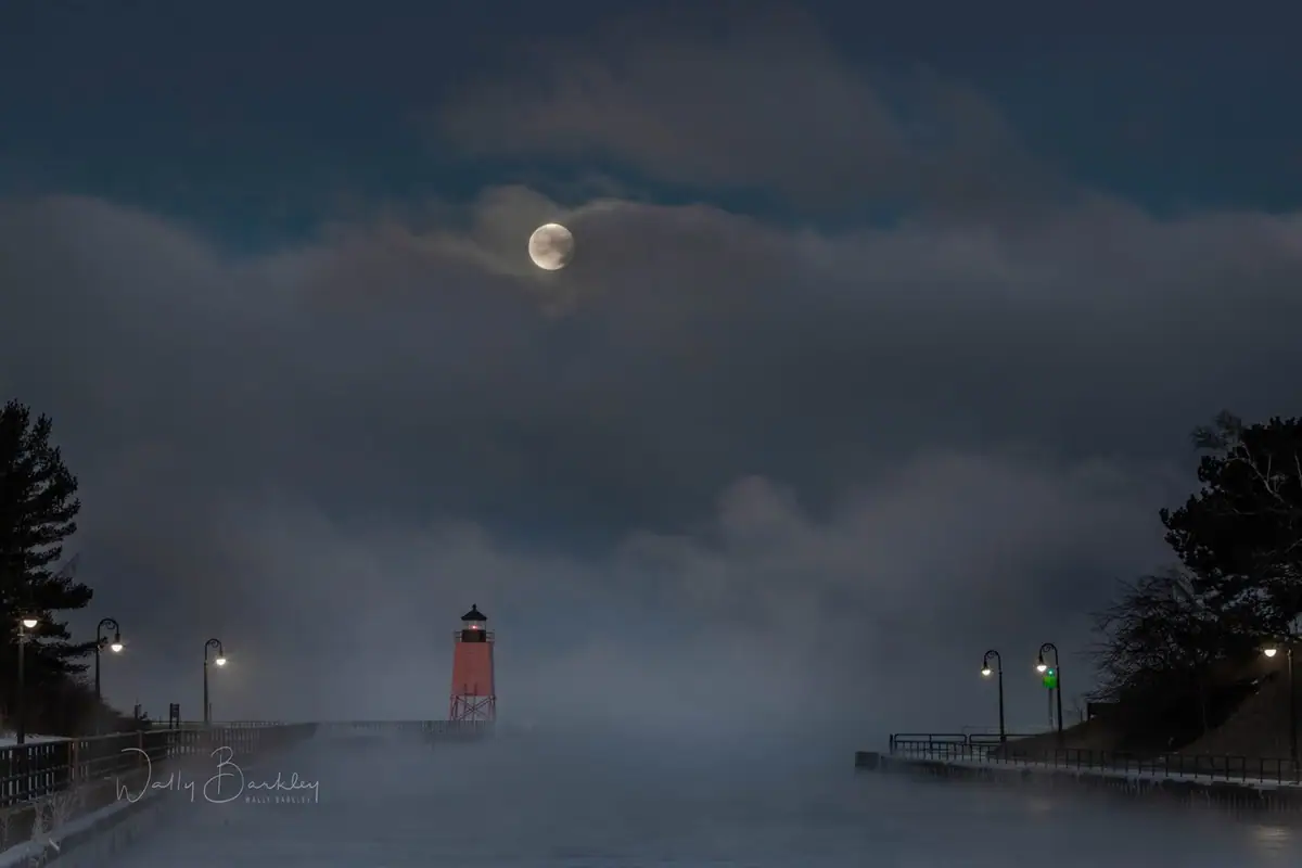A red lighthouse stands at the end of a misty pier beneath a partly veiled moon.