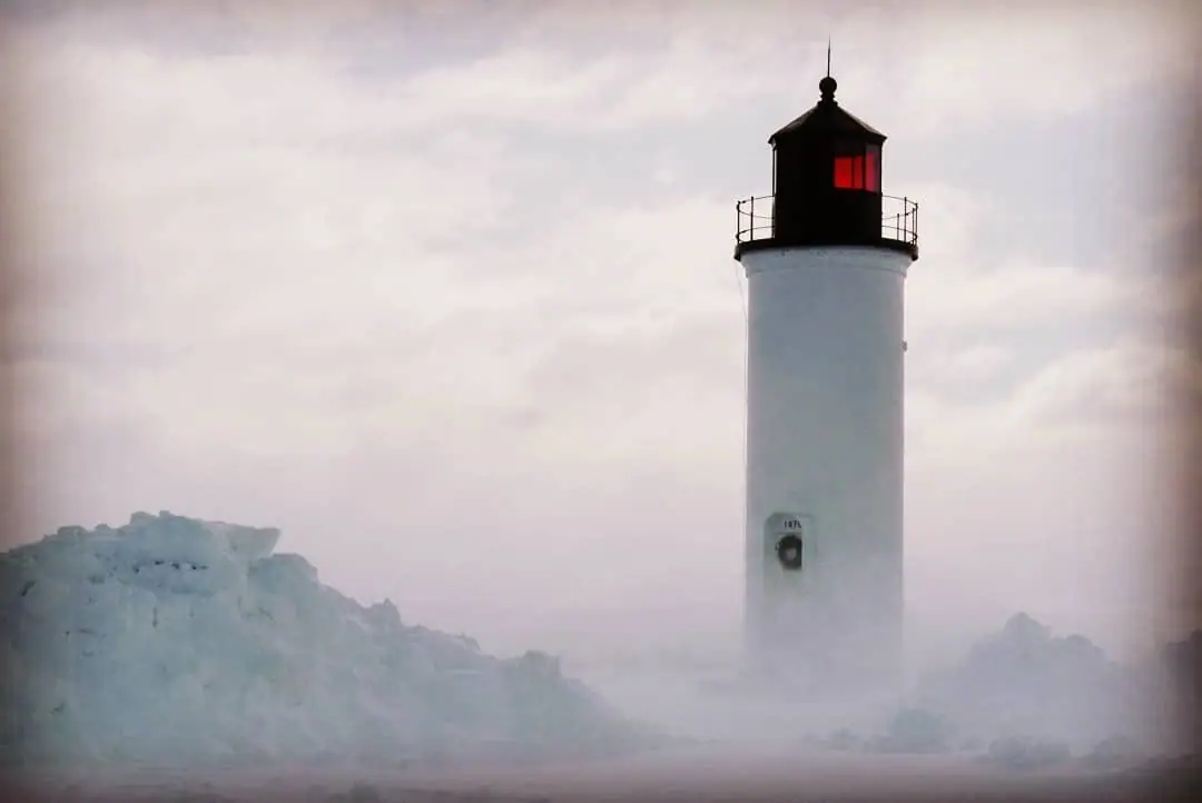 A white lighthouse rises above large snow and ice formations in a hazy winter scene.