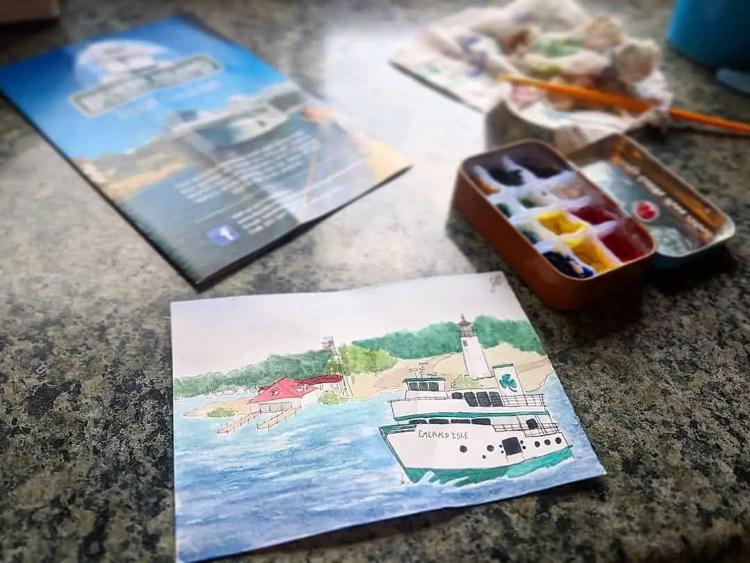A watercolor ferry painting, brochure, and open paint palette with brushes on a countertop.