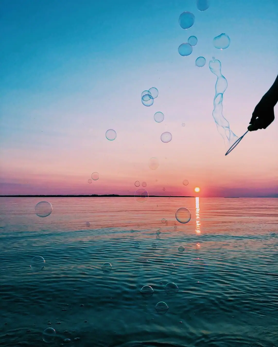 A silhouetted hand holds a bubble wand above rippling water as bubbles rise against a colorful sunset.