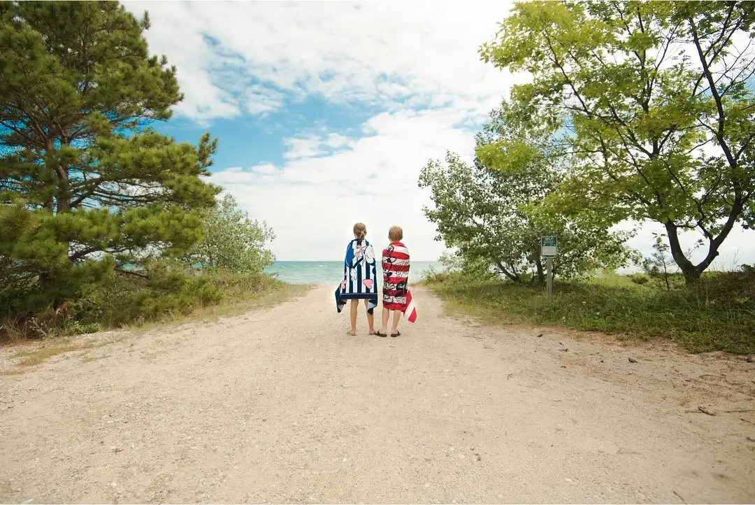 Two children wrapped in beach towels walk down a sandy path toward the water.