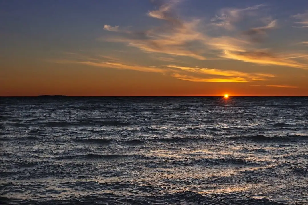 Sunset over choppy open water with a low dark shoreline on the horizon.