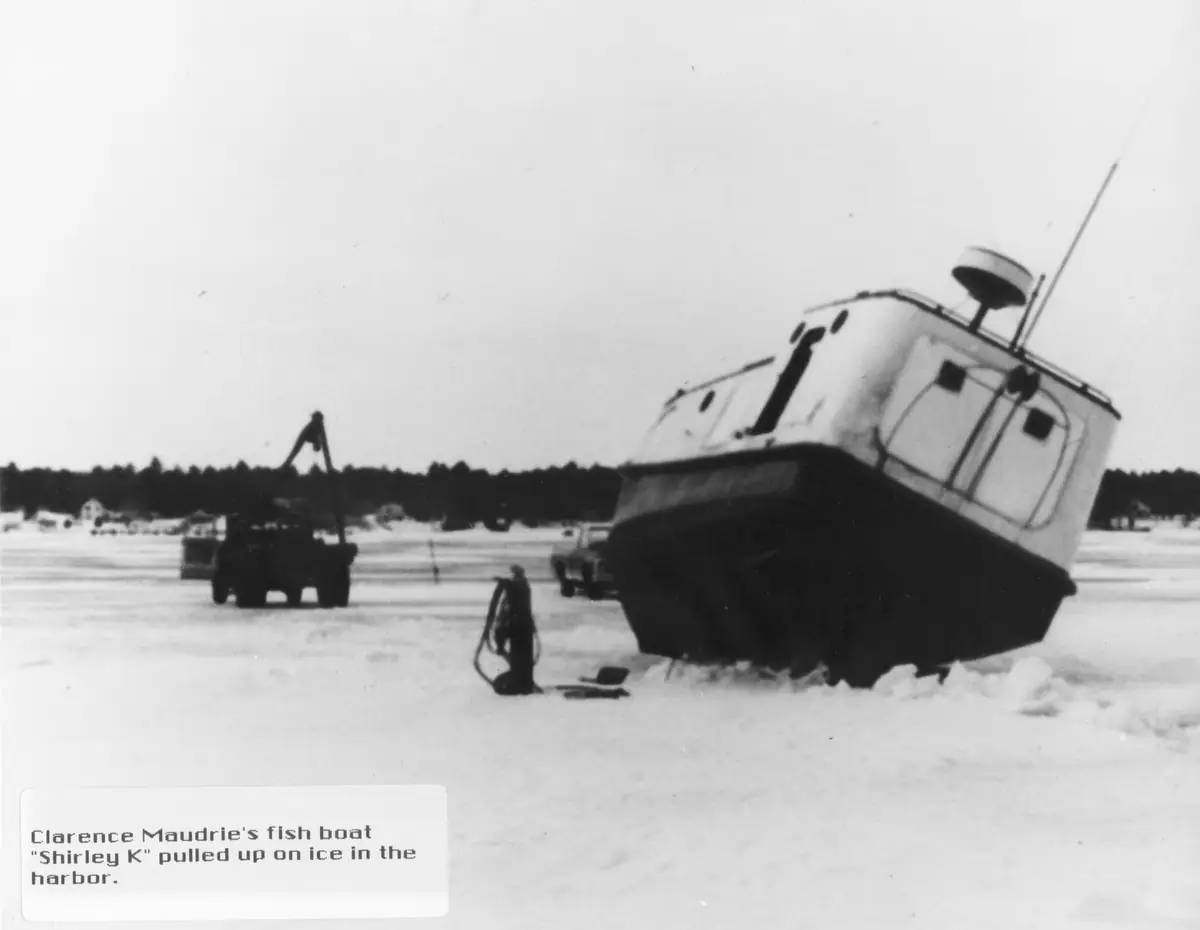 A fishing boat rests tilted on frozen water while a crane truck and person stand nearby.
