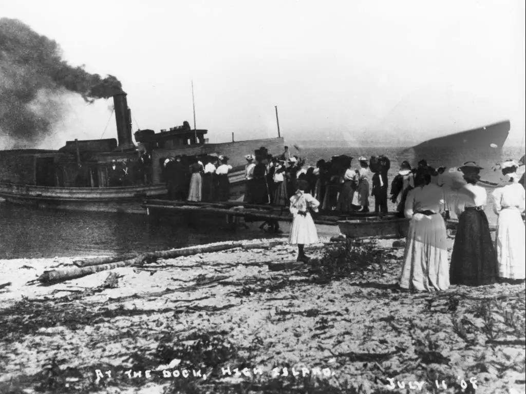 A steam-powered boat is alongside a dock while people gather on the sandy shore.