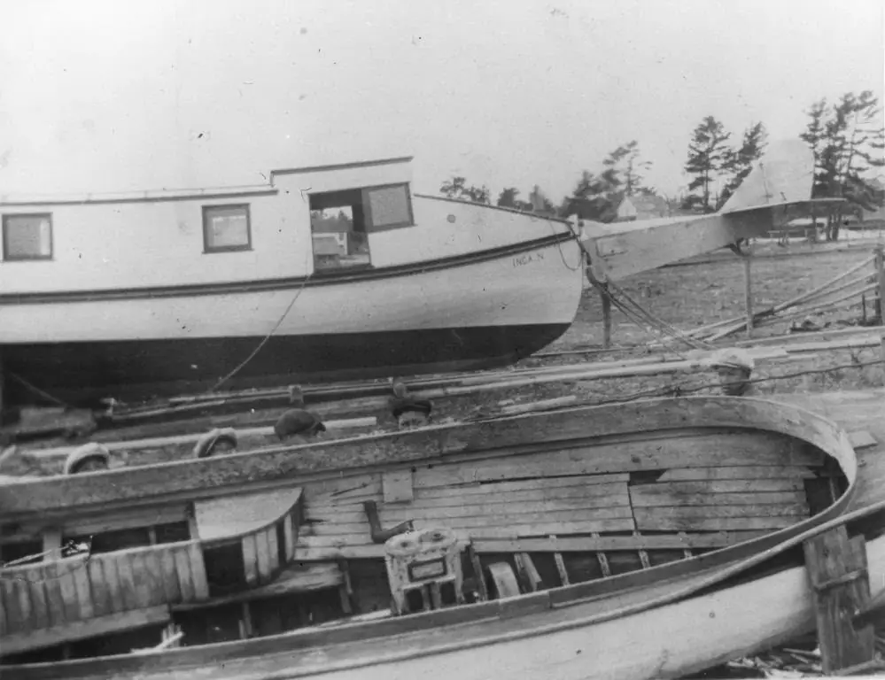 Two wooden boats rest on rails at a boatyard, with the name “INCA N” visible on the rear boat.