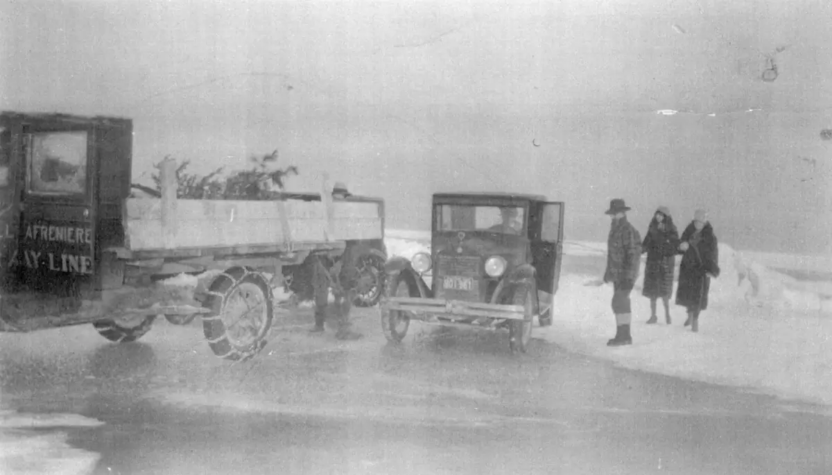 A truck, an old car, and three people on a snow-covered road in blowing winter conditions.