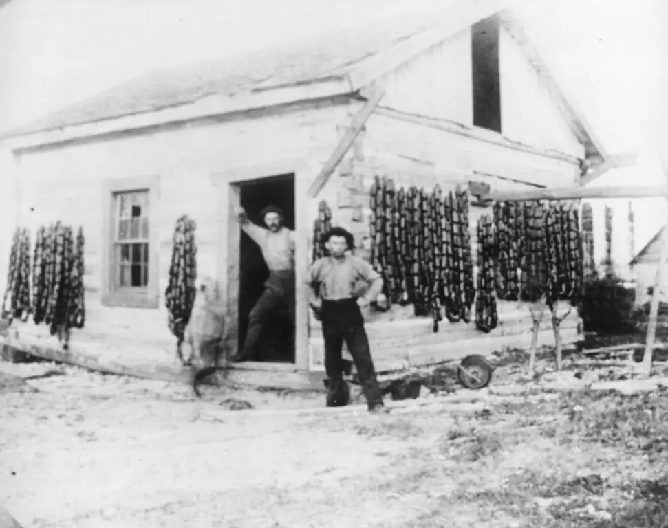 Two people and a dog stand outside a log building surrounded by rows of hanging fishing gear.