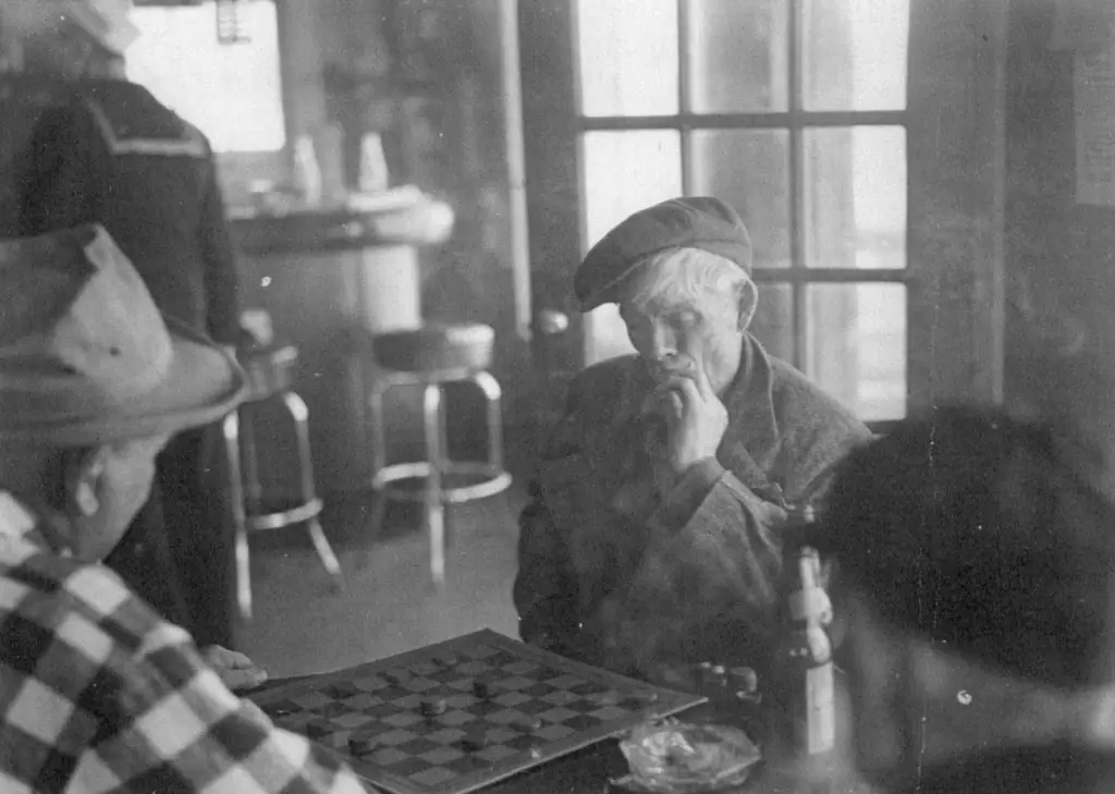 Two men play a board game at a table inside a room with large windows.