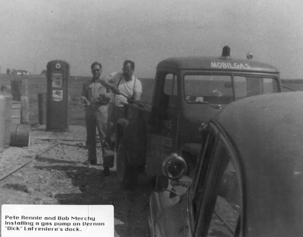 Two men stand beside a vintage fuel truck and gasoline pump on a dock.
