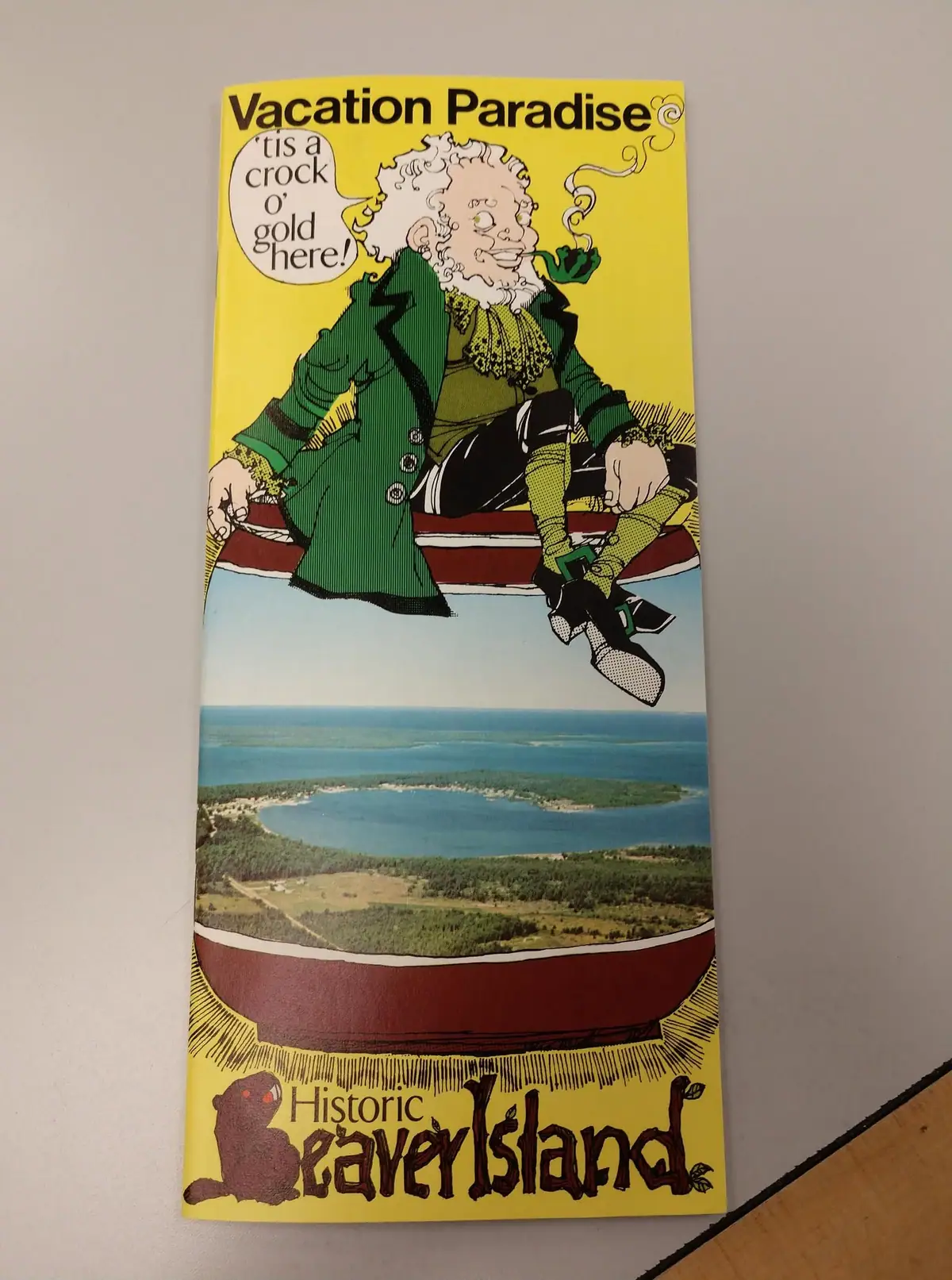Photograph of a yellow travel brochure showing a cartoon figure above an aerial landscape photograph.