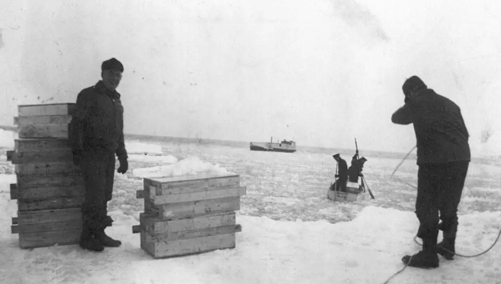 Two bundled men stand beside stacked wooden crates on a snowy, icy expanse, with a small vessel in the distance.