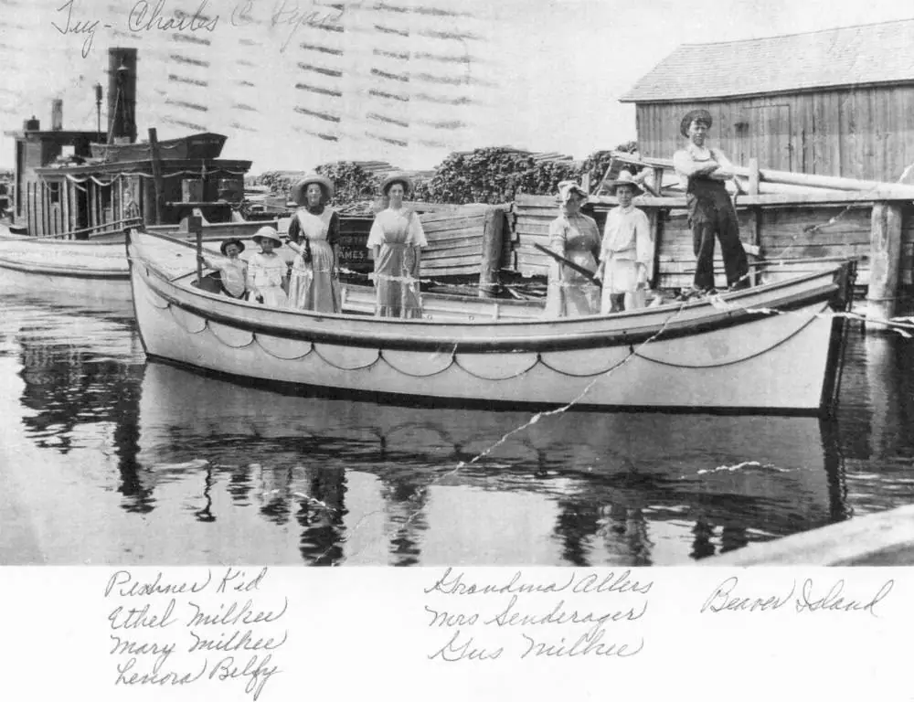 A group of women and children pose aboard a decorated boat beside a wooden dock and waterfront buildings.