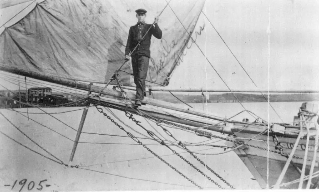 A sailor stands among the rigging and sails on the bow of a sailing vessel.