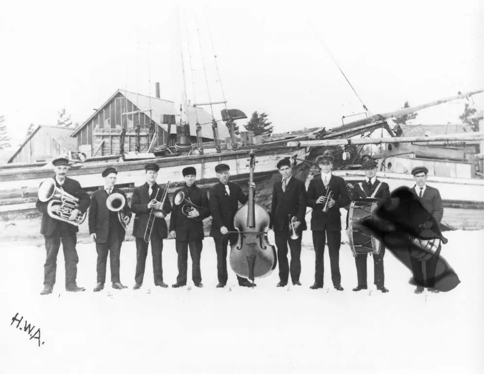 Nine men pose as a band with brass instruments, an upright bass, and a drum beside a wooden vessel.