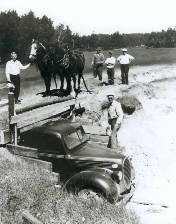 Men, horses, and an automobile stand beside a dug-out roadway and wooden planks.