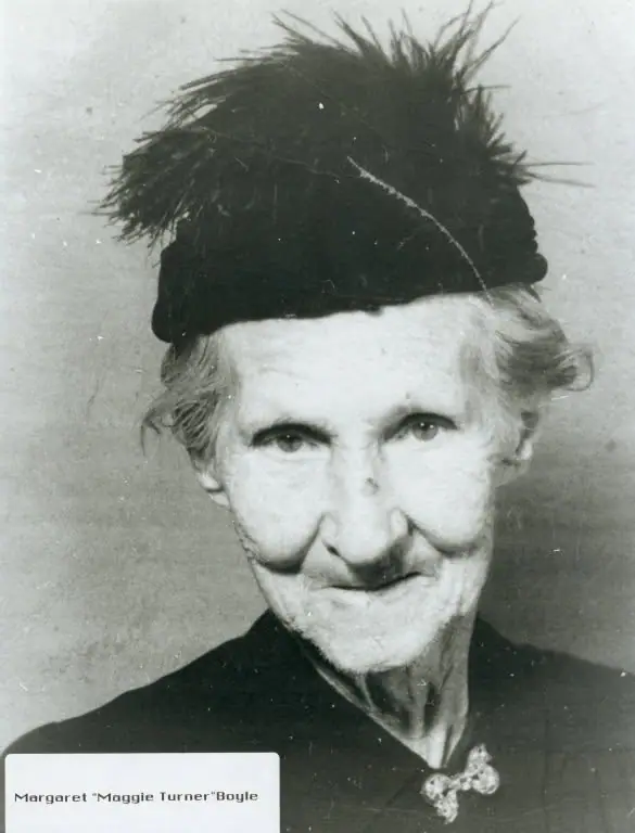Black-and-white portrait of an older woman in a feathered hat and dark clothing, with a printed name label below.