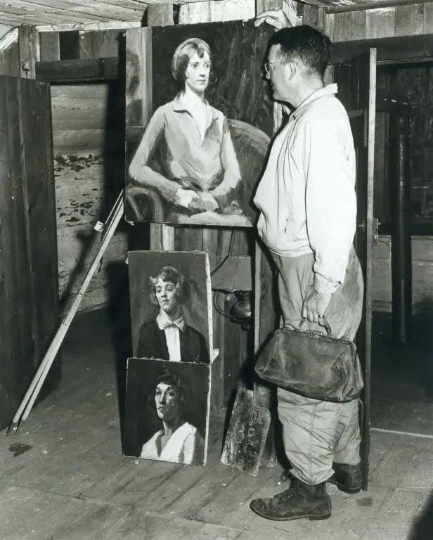 A man examines three portrait paintings in a rustic wooden interior while carrying a leather briefcase.