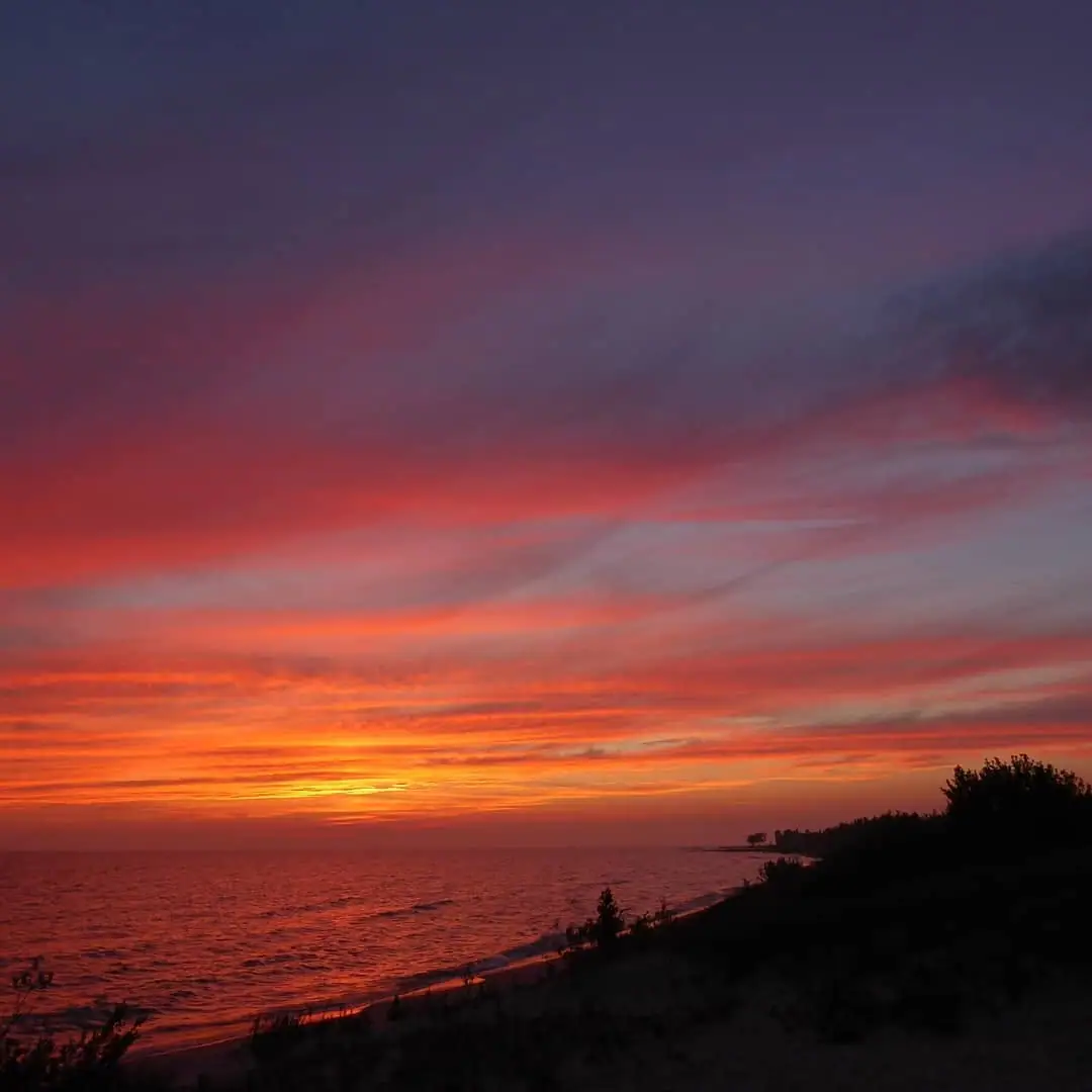 Sunset casts orange and red light across rippling water beside a dark, vegetated shoreline.