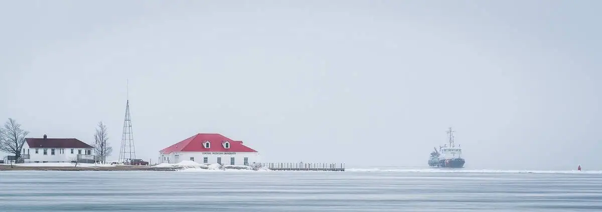 Snow-covered waterfront with buildings, a communications tower, pier, and distant vessel.