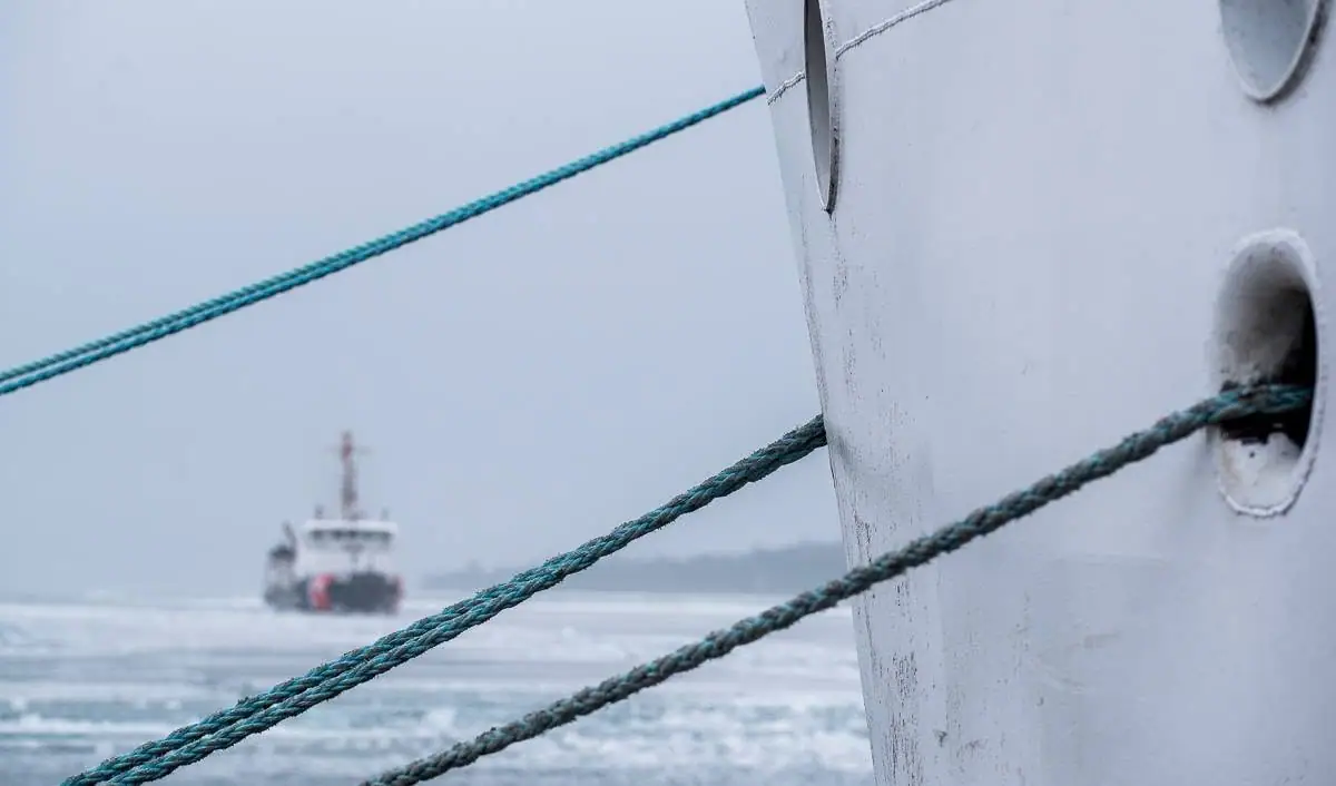 Mooring ropes cross a weathered white vessel beside icy water, with a small workboat in the distance.
