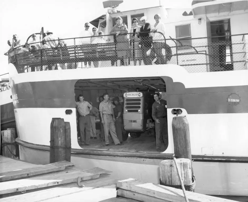 A ferry at a wooden dock with passengers above and workers beside a large vehicle in its open cargo bay.
