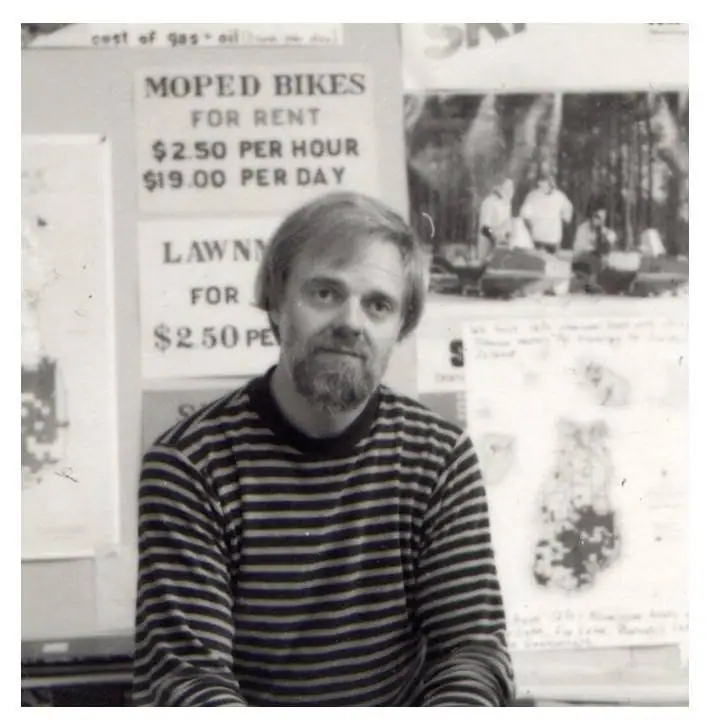 Black-and-white portrait of a bearded man seated before rental and service signs.