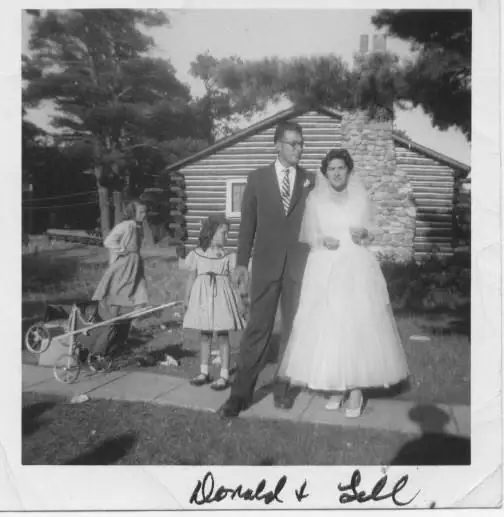 Black-and-white wedding portrait of a couple and two children before a log cabin.