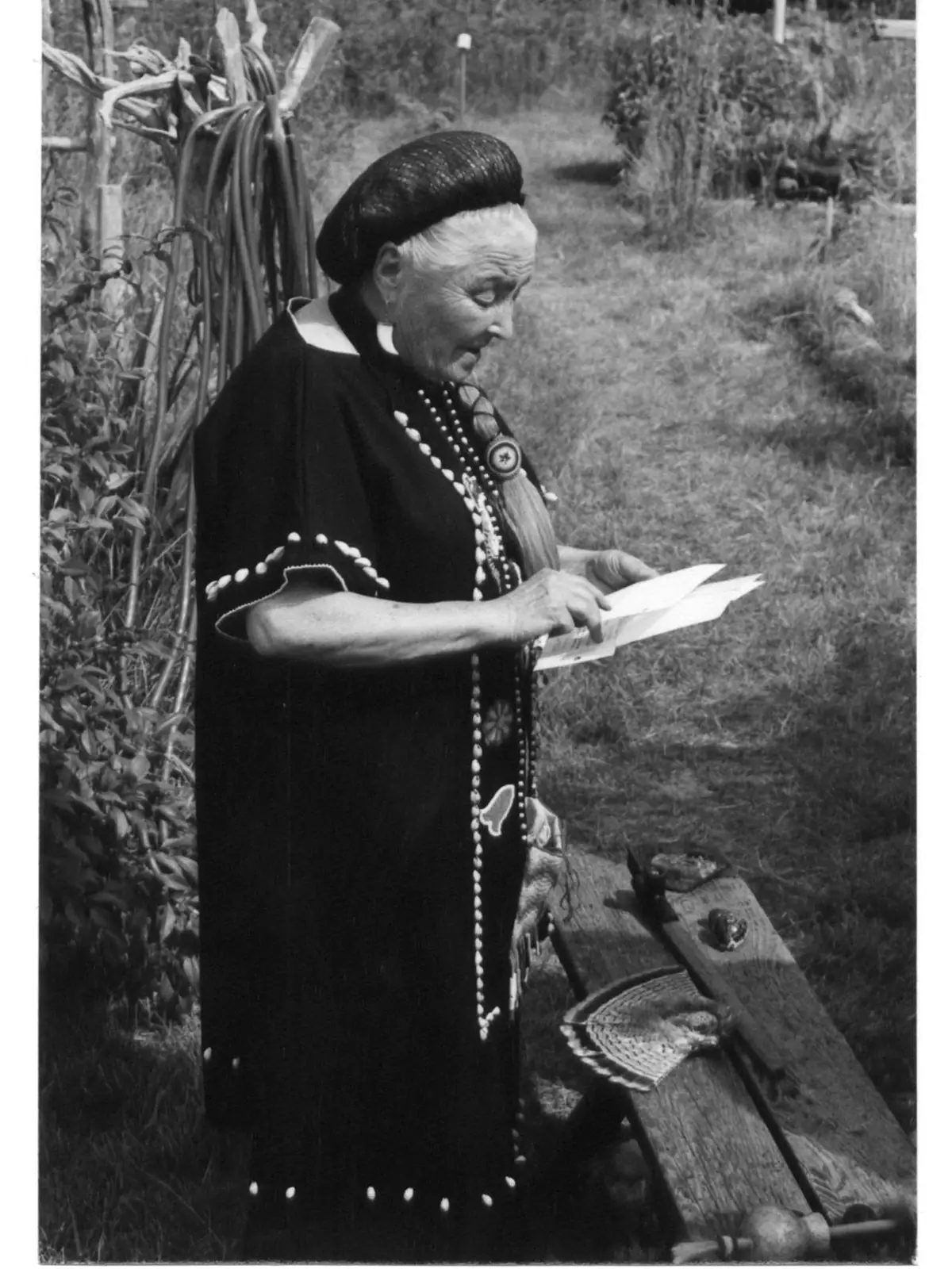 An elderly woman in a dark decorated dress reads papers beside a wooden bench in a garden.