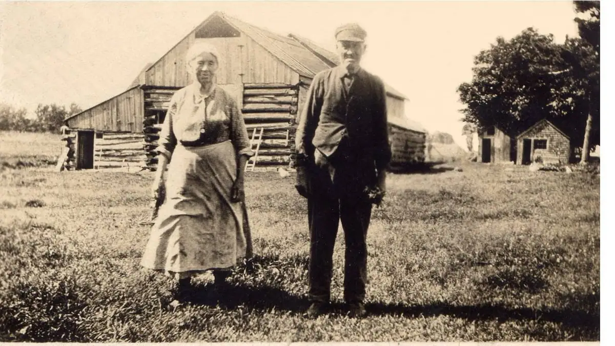 Two adults stand in a grassy yard before a large log barn and smaller outbuildings.