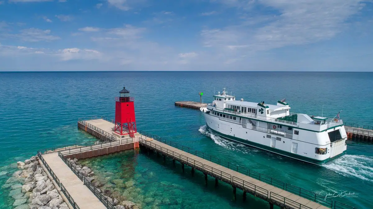 Aerial photograph of a white ferry passing a red lighthouse and piers in clear blue water.