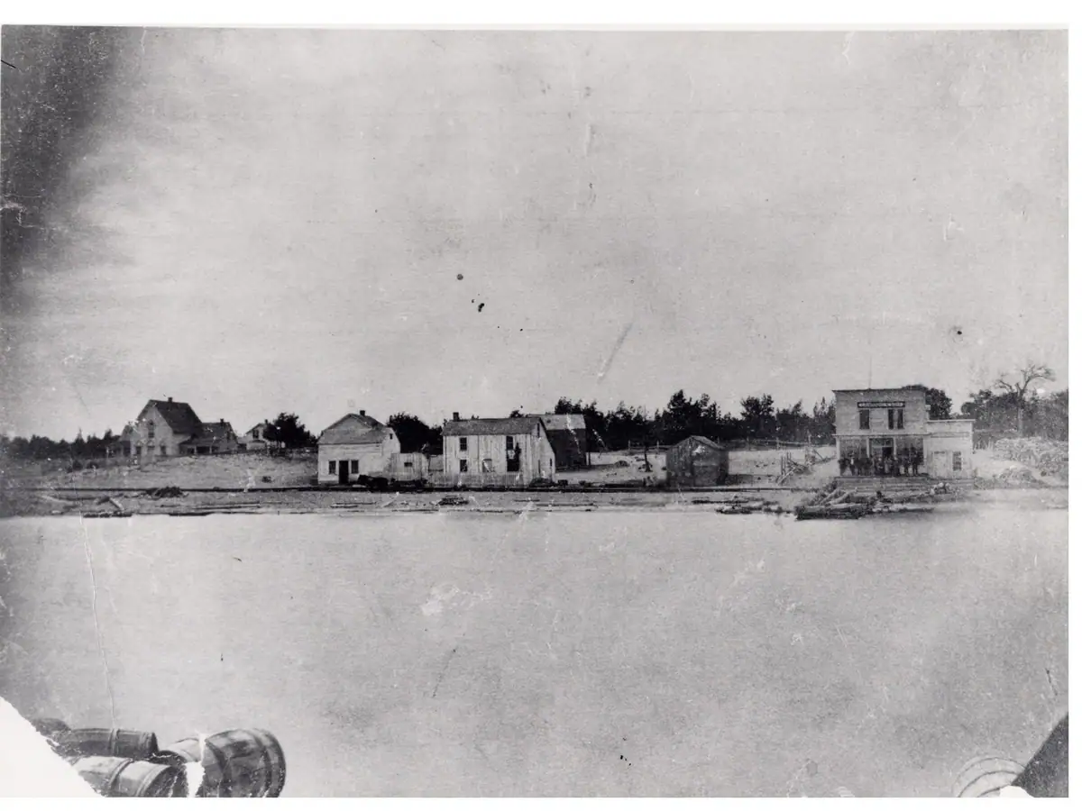 Black-and-white waterfront view of a small settlement with houses, docks, and a larger two-story building.
