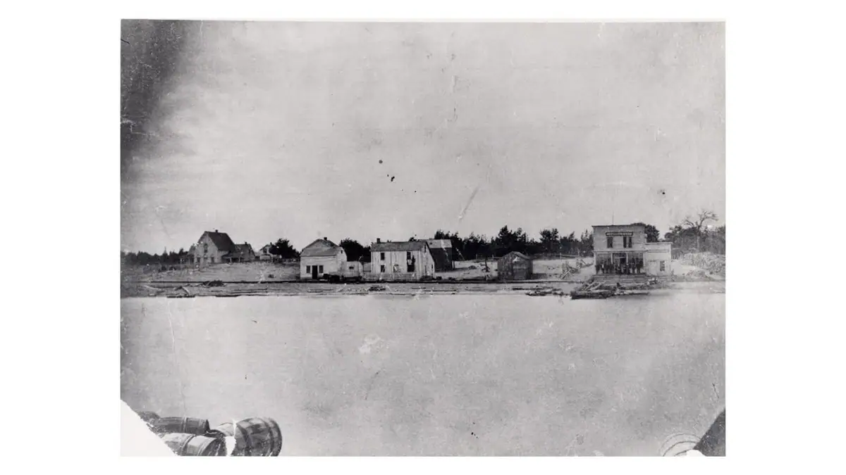 Black-and-white photograph of a waterfront settlement with houses, a prominent commercial building, and barrels in the foreground.