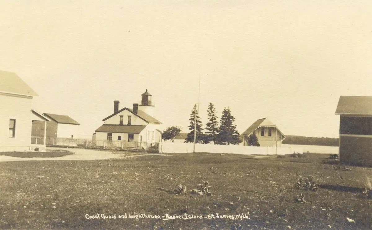 Sepia photograph of a lighthouse complex with surrounding buildings, fence, trees, and a distant shoreline.