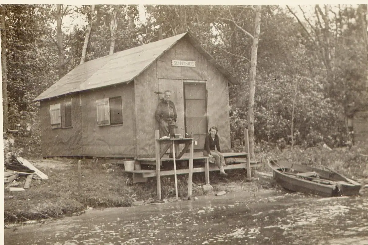 Two adults pose beside a small wooden building marked “SUNNYSIDE,” with a narrow boat in the foreground.