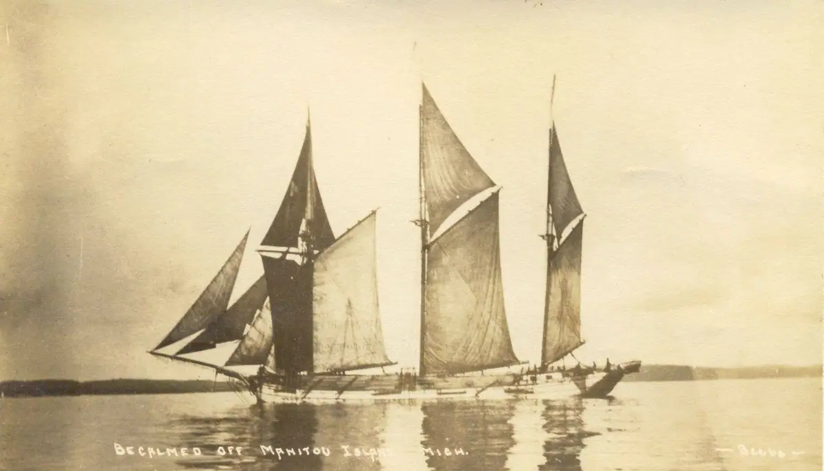 Sepia photograph of three sailing vessels becalmed on calm water before a low shoreline.