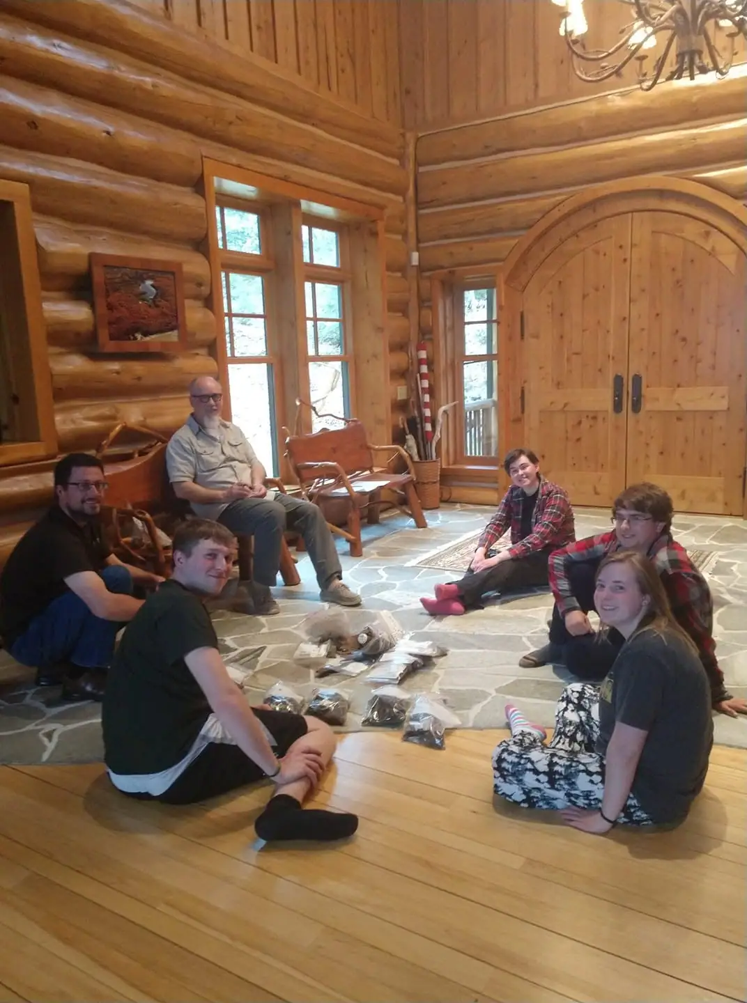 Six people sit around bagged objects on the floor inside a log-built room.