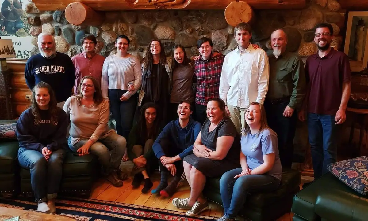 Fifteen people pose in two rows inside a rustic room with a stone wall, log beams, sofas, and a patterned rug.