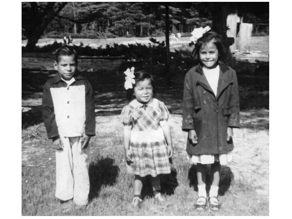 Three children stand outdoors on a grassy lawn, wearing jackets and dresses with large hair bows.