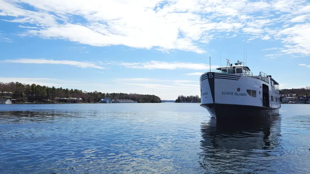 A white ferry approaches across calm water with wooded shoreline and buildings behind it.