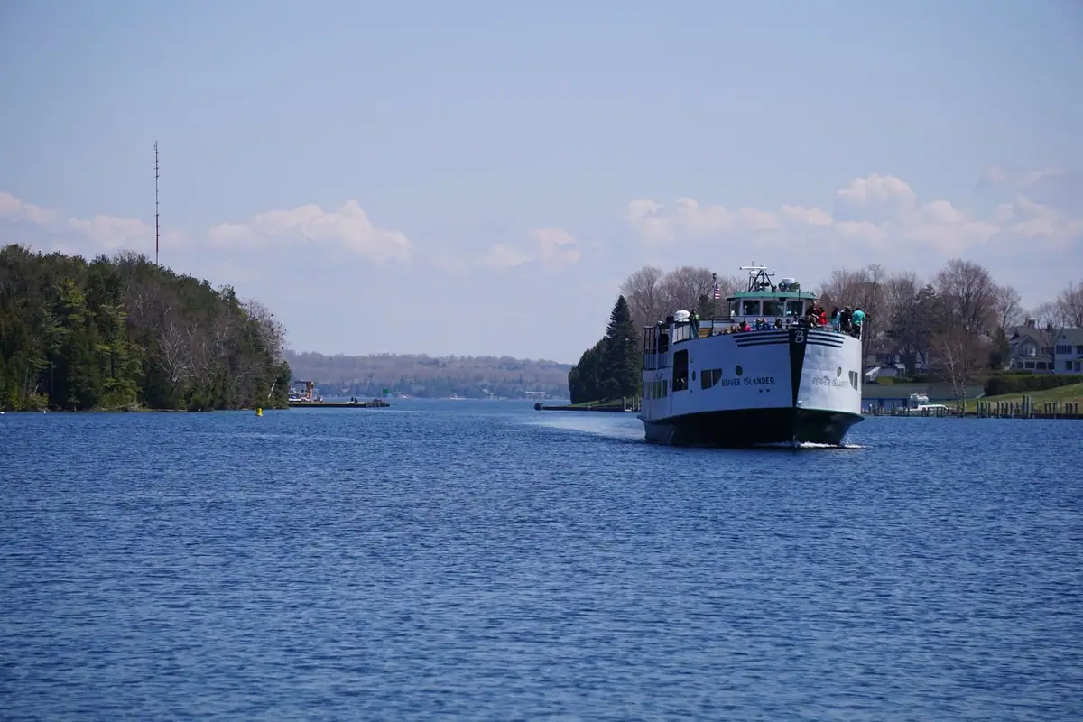 A passenger ferry approaches along a blue waterway between wooded and residential shores.