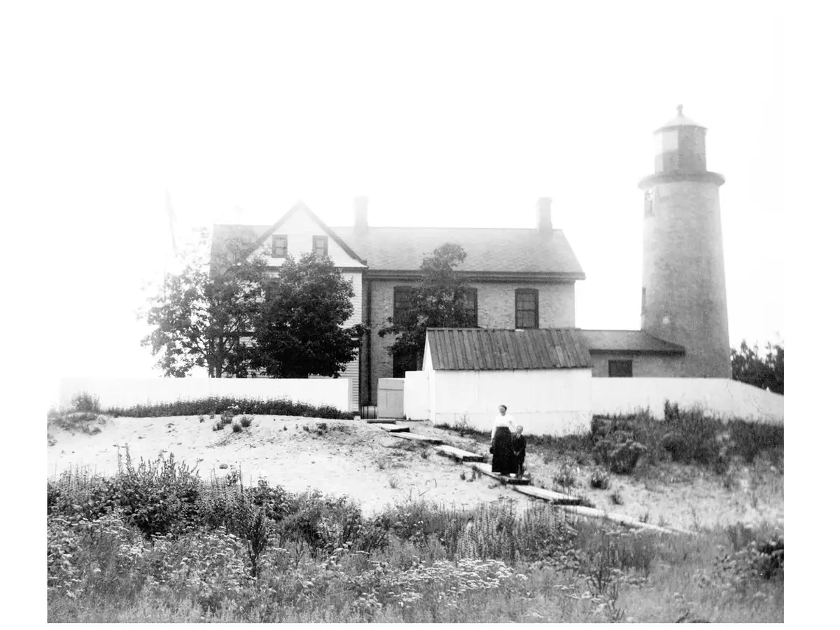 Black-and-white photograph of a lighthouse, adjoining buildings, and two people on a sandy path.