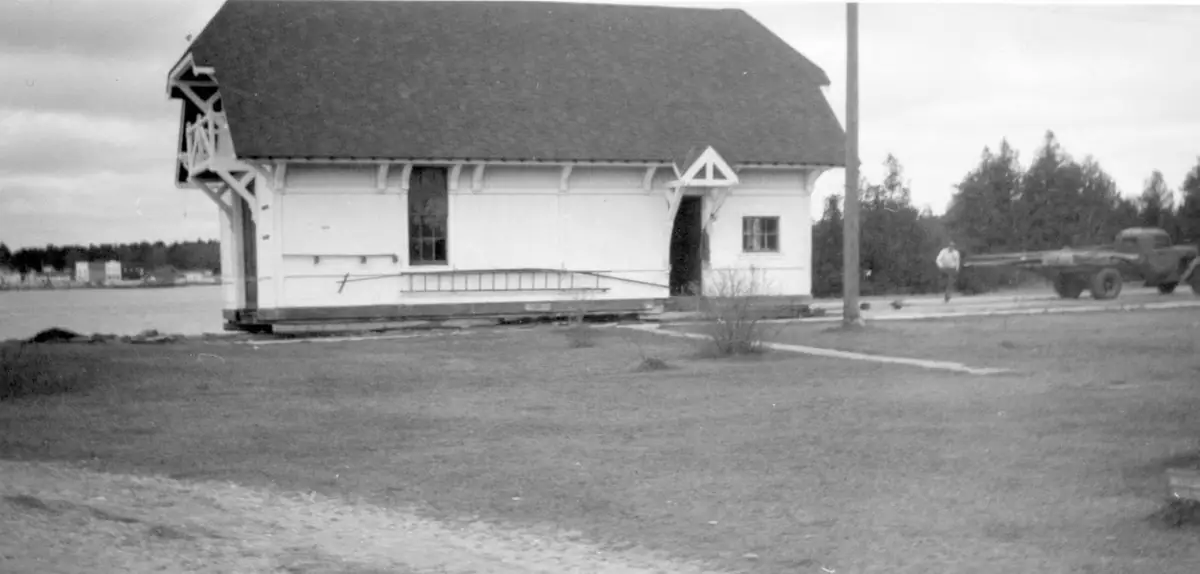 Black-and-white photograph of a wooden gabled building beside the water, with a truck and pedestrian nearby.