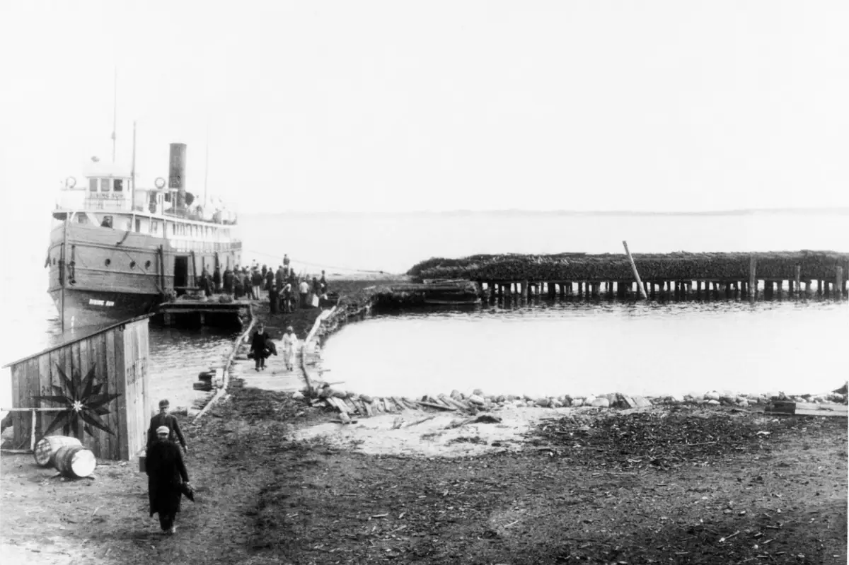 A passenger steamer is docked beside a wooden pier while people walk along the shore.