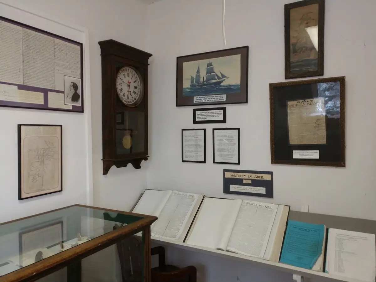 Museum display featuring framed documents, open newspapers, a wall clock, and a sailing ship illustration.