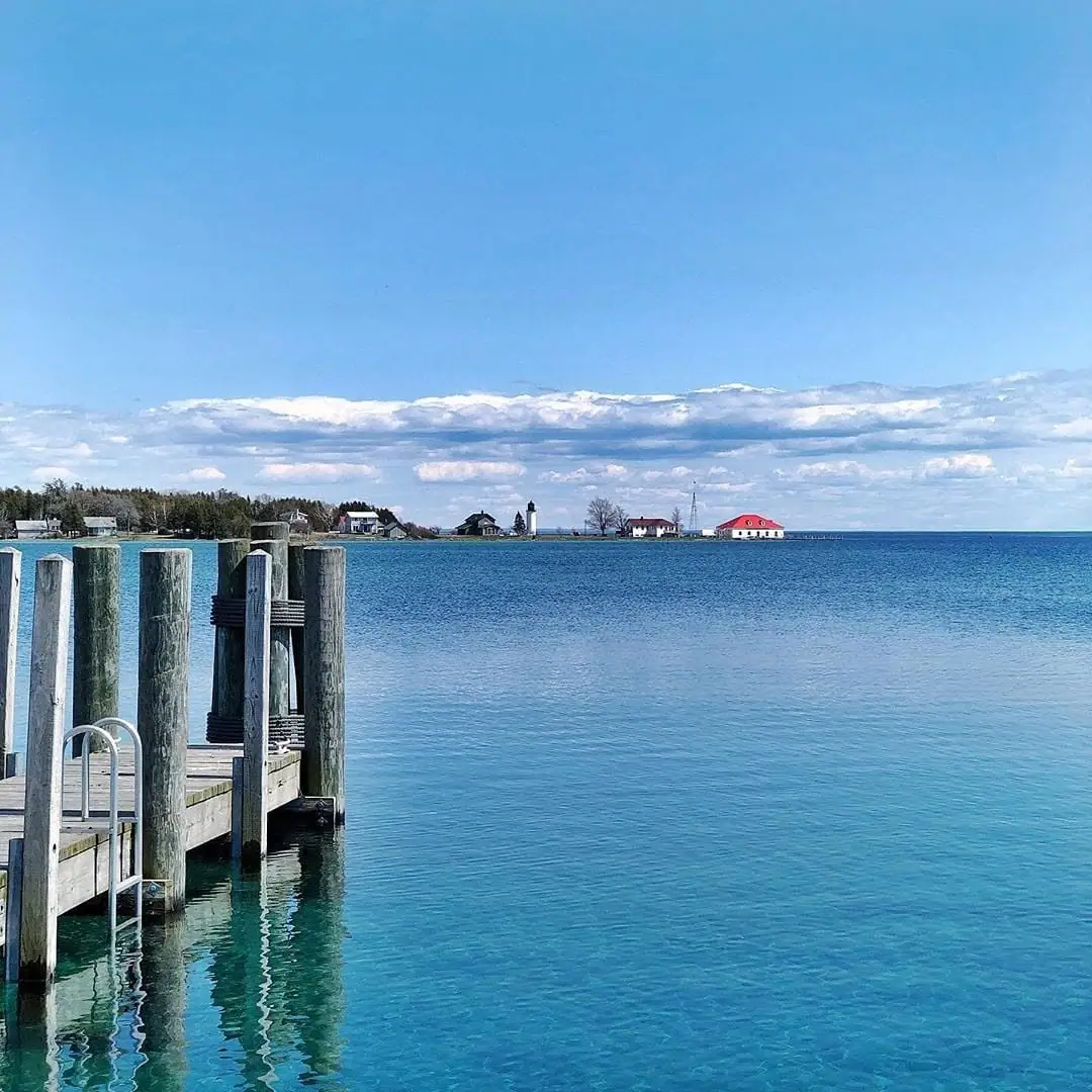A wooden dock extends across bright blue water toward a distant shoreline with houses and a lighthouse.